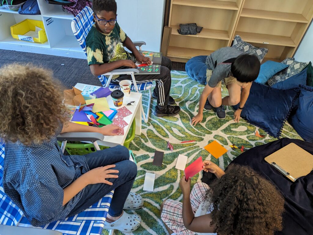 Four students gathered around table and bean bags, folding origami.
